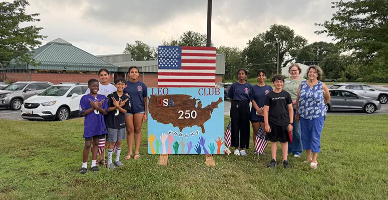 Group of children and adults posing beside a handmade sign reading LEO Club USA 250, with an American flag overhead.