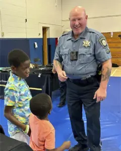 Police officer in uniform talking with two young boys in a gym with blue mats and a table nearby, a friendly interaction.