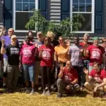 Group of volunteers posing outside a gray building, many waving at the camera. (Informative group photo)