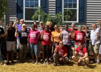 Group of volunteers posing outside a gray building, many waving at the camera. (Informative group photo)