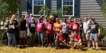 Group of volunteers posing outside a gray building, many waving at the camera. (Informative group photo)