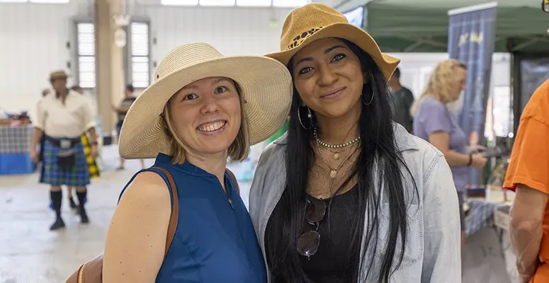 Two smiling women in wide-brim hats posing together at an indoor community event.