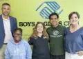 Group of five smiling people standing together in front of a Boys & Girls Clubs sign and logo (two adults on ends, three youths in the center).