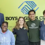 Group of five smiling people standing together in front of a Boys & Girls Clubs sign and logo (two adults on ends, three youths in the center).