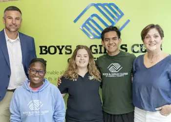 Group of five smiling people standing together in front of a Boys & Girls Clubs sign and logo (two adults on ends, three youths in the center).