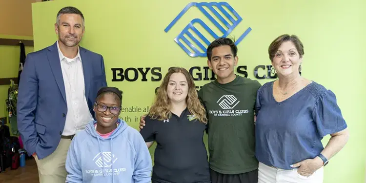 Group of five smiling people standing together in front of a Boys & Girls Clubs sign and logo (two adults on ends, three youths in the center).