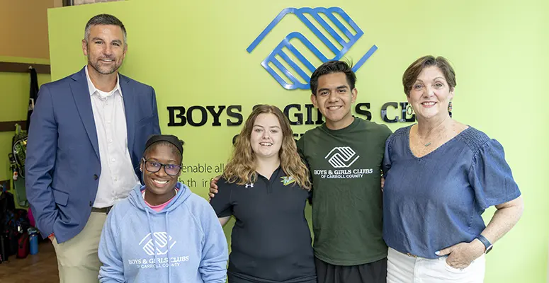 Group of five smiling people standing together in front of a Boys & Girls Clubs sign and logo (two adults on ends, three youths in the center).