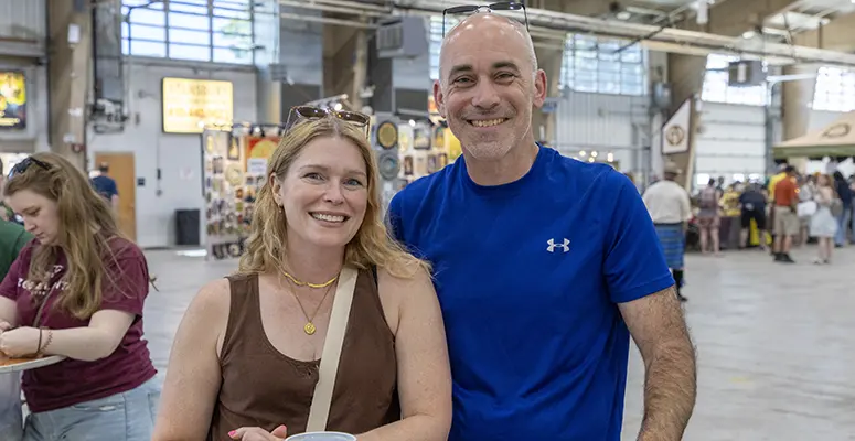 Smiling couple posing together at an indoor event, the man in a blue athletic shirt and the woman in a brown top with sunglasses on her head, with booths in the background.