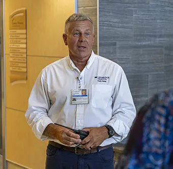 Middle-aged man in a white collared shirt with a name badge, holding glasses, standing in a hallway.