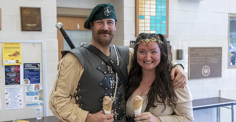 Man in medieval armor and green beret posing with a smiling woman, both holding ice cream cones indoors at a community hall.
