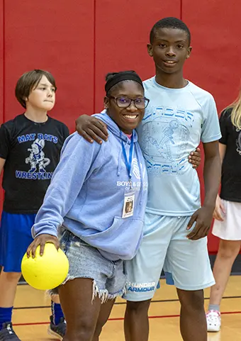 Smiling girl with glasses in a light blue hoodie and denim shorts holds a yellow ball, posing with a taller boy in a gym.