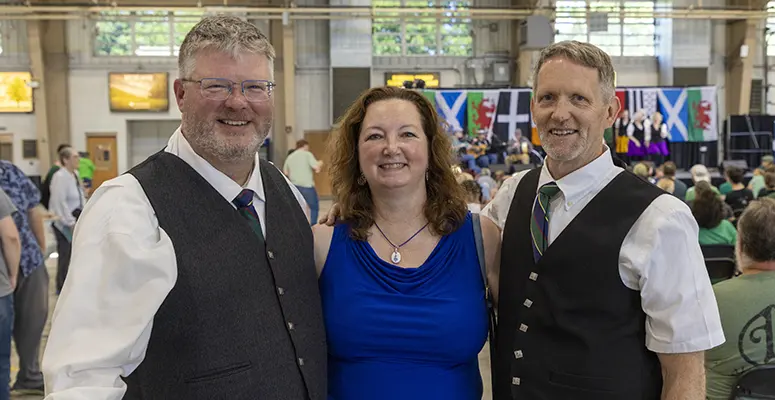 Three adults posing at an indoor event; two men in vests flank a woman in a blue dress, with flags and attendees behind them.