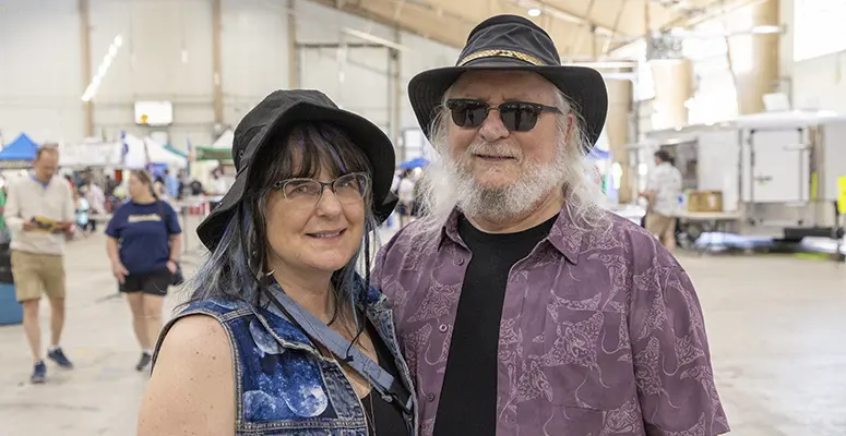 Smiling woman in a denim vest and hat beside a bearded man in a purple shirt and black hat at an indoor event.