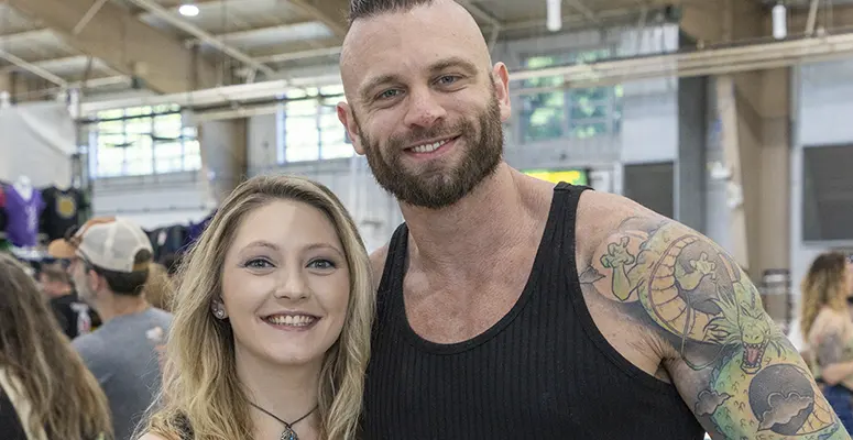 Smiling man with a tattooed left arm stands beside a smiling blonde woman at a crowded indoor event venue.