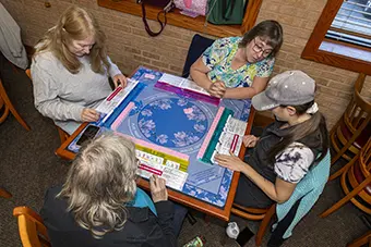 Four people sit around a table playing a board game with a blue circular board and tiles in a cozy room.