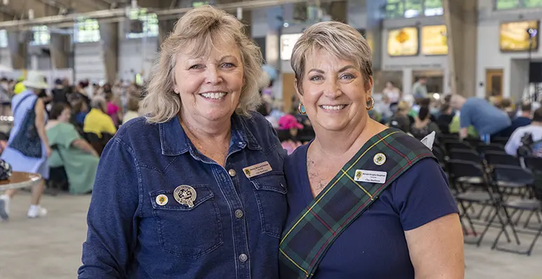 Two smiling women at a busy indoor event pose for a photo, wearing name badges.