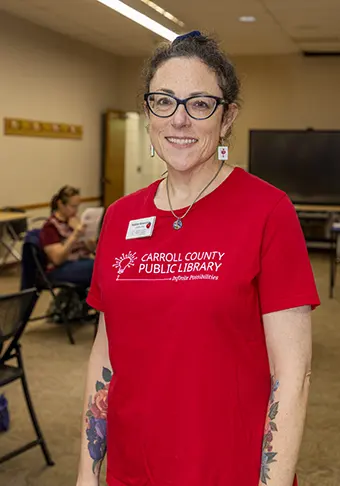 Smiling woman wearing a red Carroll County Public Library polo with name tag, standing in a classroom-like room with people in the background.