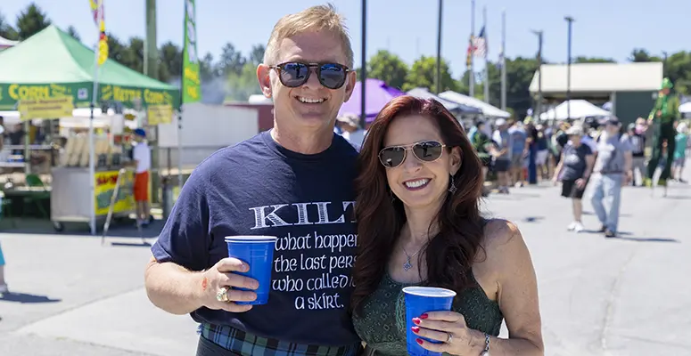 Smiling couple wearing sunglasses at an outdoor festival, each holding a blue cup.