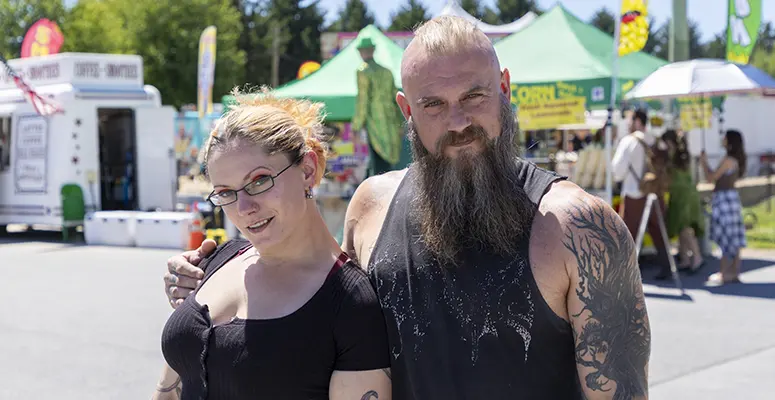 Couple posing together at an outdoor festival with colorful tents and stalls in the background.