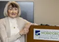 Smiling older woman stands at a podium with a Carroll County Workforce Development sign in a conference room.
