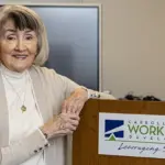 Smiling older woman stands at a podium with a Carroll County Workforce Development sign in a conference room.