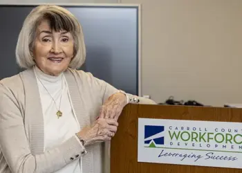 Smiling older woman stands at a podium with a Carroll County Workforce Development sign in a conference room.