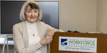 Smiling older woman stands at a podium with a Carroll County Workforce Development sign in a conference room.