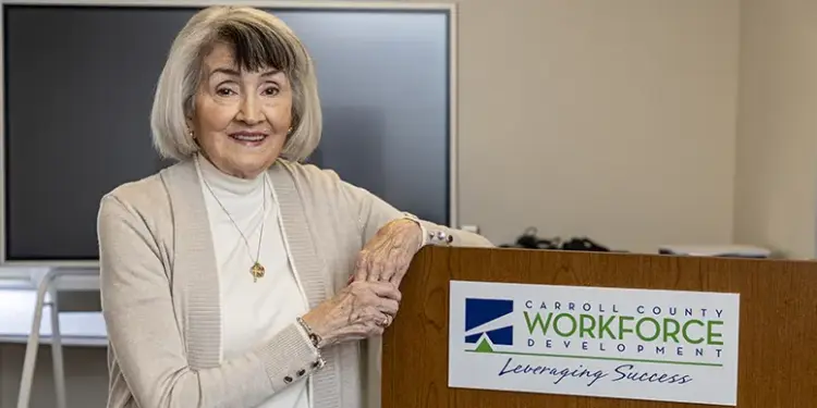 Smiling older woman stands at a podium with a Carroll County Workforce Development sign in a conference room.