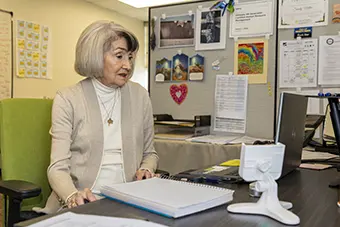 Older woman with short blond hair sits at a desk in a bright office, reviewing documents beside a laptop.