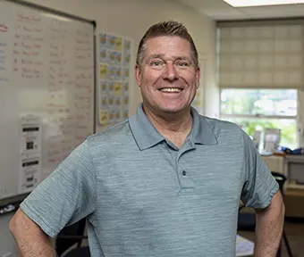 Smiling man in a blue polo shirt standing in a classroom with a whiteboard and posters in the background.