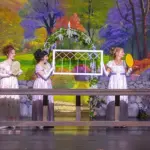 Five women in vintage white dresses stand behind a long wooden table on a theater stage, each holding a handheld fan or tambourine, with a colorful garden backdrop and a white lattice arch centerpiece.