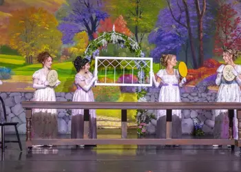 Five women in vintage white dresses stand behind a long wooden table on a theater stage, each holding a handheld fan or tambourine, with a colorful garden backdrop and a white lattice arch centerpiece.