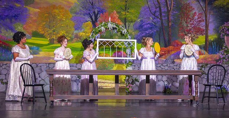 Five women in vintage white dresses stand behind a long wooden table on a theater stage, each holding a handheld fan or tambourine, with a colorful garden backdrop and a white lattice arch centerpiece.