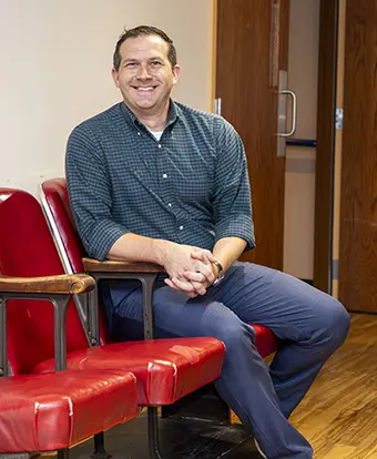 Stephen Strosnider_headshot - Carroll News | Carroll Publications | Carroll County, MD Smiling man in a teal checkered shirt and dark blue pants sits on red vinyl chairs in a waiting room, posing for the photo.
