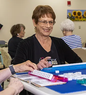 Smiling elderly woman wearing glasses sits at a table, playing a game with bingo cards and chips in a social setting.