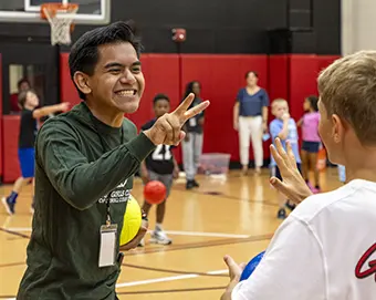 Smiling young man in a gym makes a peace sign at a youth basketball event, with kids playing in the background.