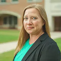 Portrait of a woman with long blonde hair, wearing a teal blouse and black blazer, standing outdoors in front of a brick building.