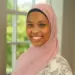 Portrait of a smiling woman wearing a pink hijab indoors by a window with greenery outside.