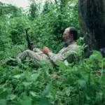 Bearded man in a light shirt sits resting against a tree in dense green forest, holding a small dark object in his hands.