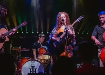 Live band on stage: a female guitarist/vocalist center stage with two guitarists and a cellist, drummer in back, colorful stage lights behind them