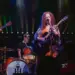 Live band on stage: a female guitarist/vocalist center stage with two guitarists and a cellist, drummer in back, colorful stage lights behind them