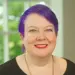 Head-and-shoulders portrait of a smiling woman with purple hair, wearing a black top, indoors by a window.