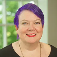 Head-and-shoulders portrait of a smiling woman with purple hair, wearing a black top, indoors by a window.