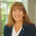 Profile photo of a smiling businesswoman with light brown hair, wearing a navy blazer and white blouse in an office setting.
