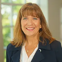 Profile photo of a smiling businesswoman with light brown hair, wearing a navy blazer and white blouse in an office setting.