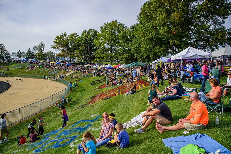 Crowd seated on a grassy hill by a curved running track, with colorful tents set up along the edge of the field and trees in the background.