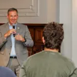 Man in a gray suit speaks into a microphone to a small audience in a conference room