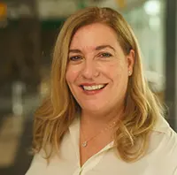 Smiling blonde woman in a white blouse, a professional headshot with an office background.