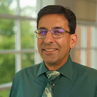 Smiling man in a green shirt and patterned tie, standing indoors near a window.