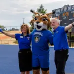 Mascot in a blue uniform with two staff members on a blue track, arms raised with pom-poms; scoreboard in the background reads 'Home of the Lynx'.